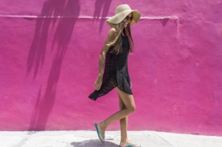 Frau ist im schwarzen Sommerkleid und Strohhut vor einer pinken Hausfassade in den Ferien